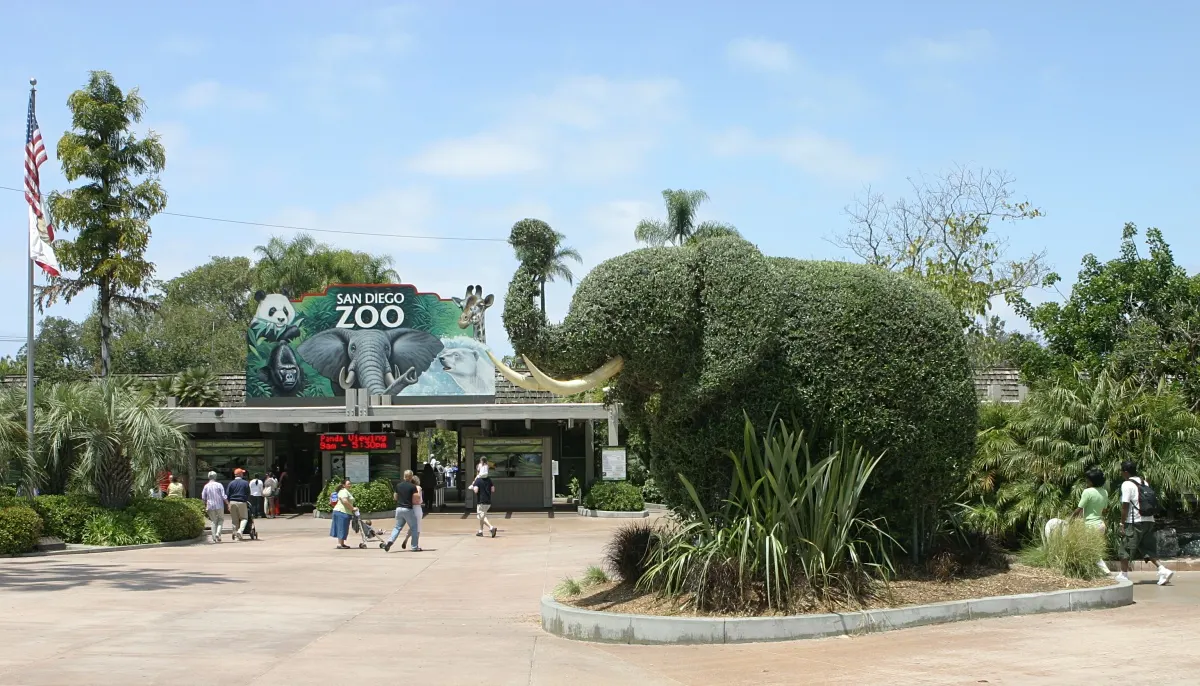 Entrada do zoológico com letreiro «San Diego Zoo» e um elefante de arbusto podado ao lado, visitantes caminhando na praça