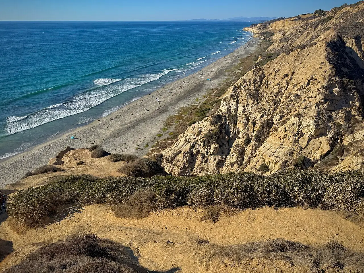Falésias de arenito amarelado e vegetação rasteira sobre uma praia longa e o mar azul