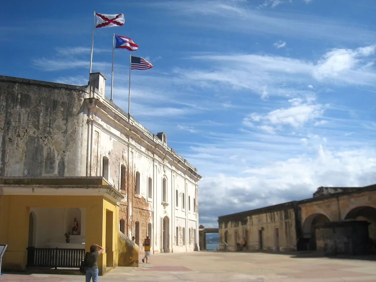 Pátio do Castillo San Cristóbal, com um edifício de pedra caiada encimado por três bandeiras, uma capelinha amarela à esquerda e arcadas ao fundo