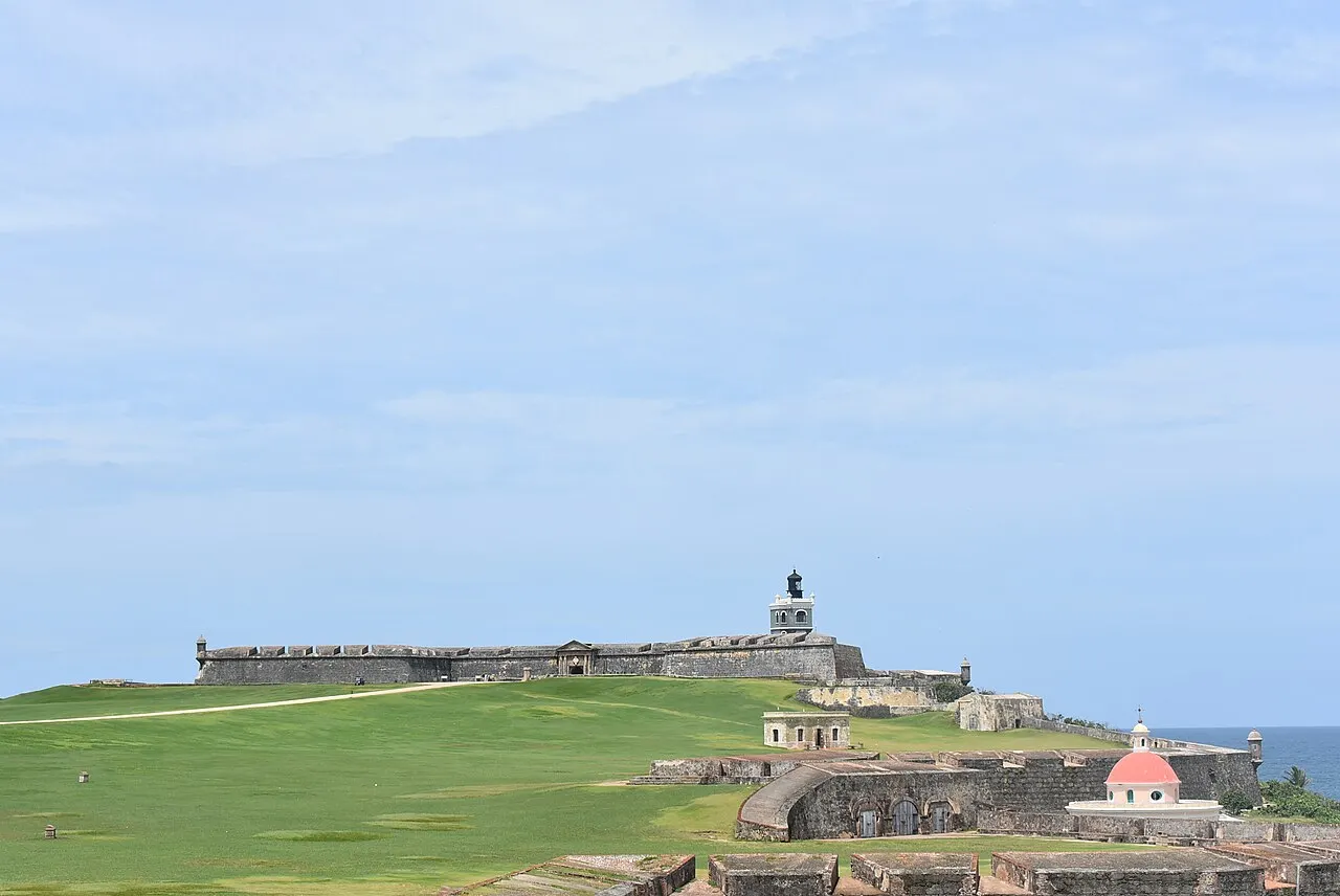 As muralhas baixas do Castillo San Felipe del Morro com o farol, no alto de um grande gramado verde, e o mar à direita sob céu claro