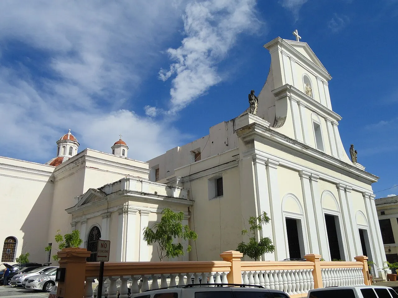Fachada branca da Catedral de San Juan Bautista, com frontão de volutas, estátuas e pequenas cúpulas laterais, atrás de uma balaustrada sob céu azul