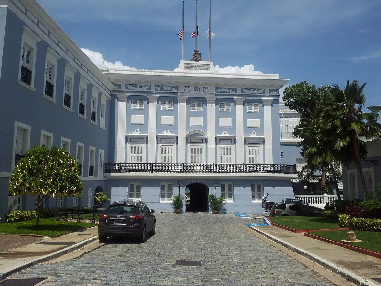 Fachada azul-clara e branca de La Fortaleza, com varanda de ferro e portão em arco, ao fim de um acesso de paralelepípedos com um carro estacionado