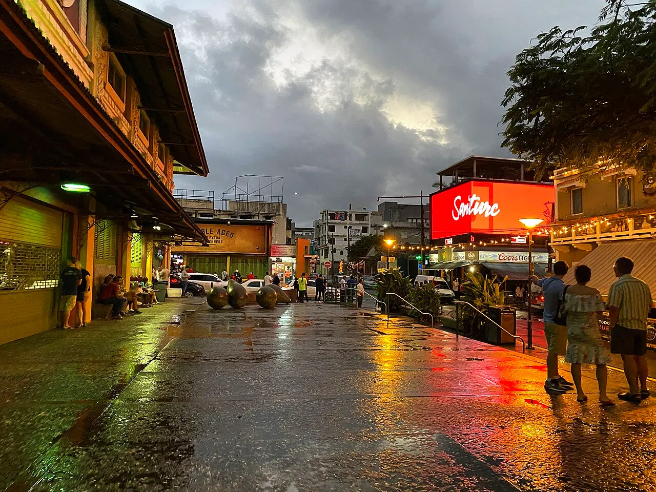 La Placita de Santurce ao anoitecer, com o calçamento molhado refletindo luzes, o mercado coberto à esquerda e um letreiro vermelho iluminado à direita