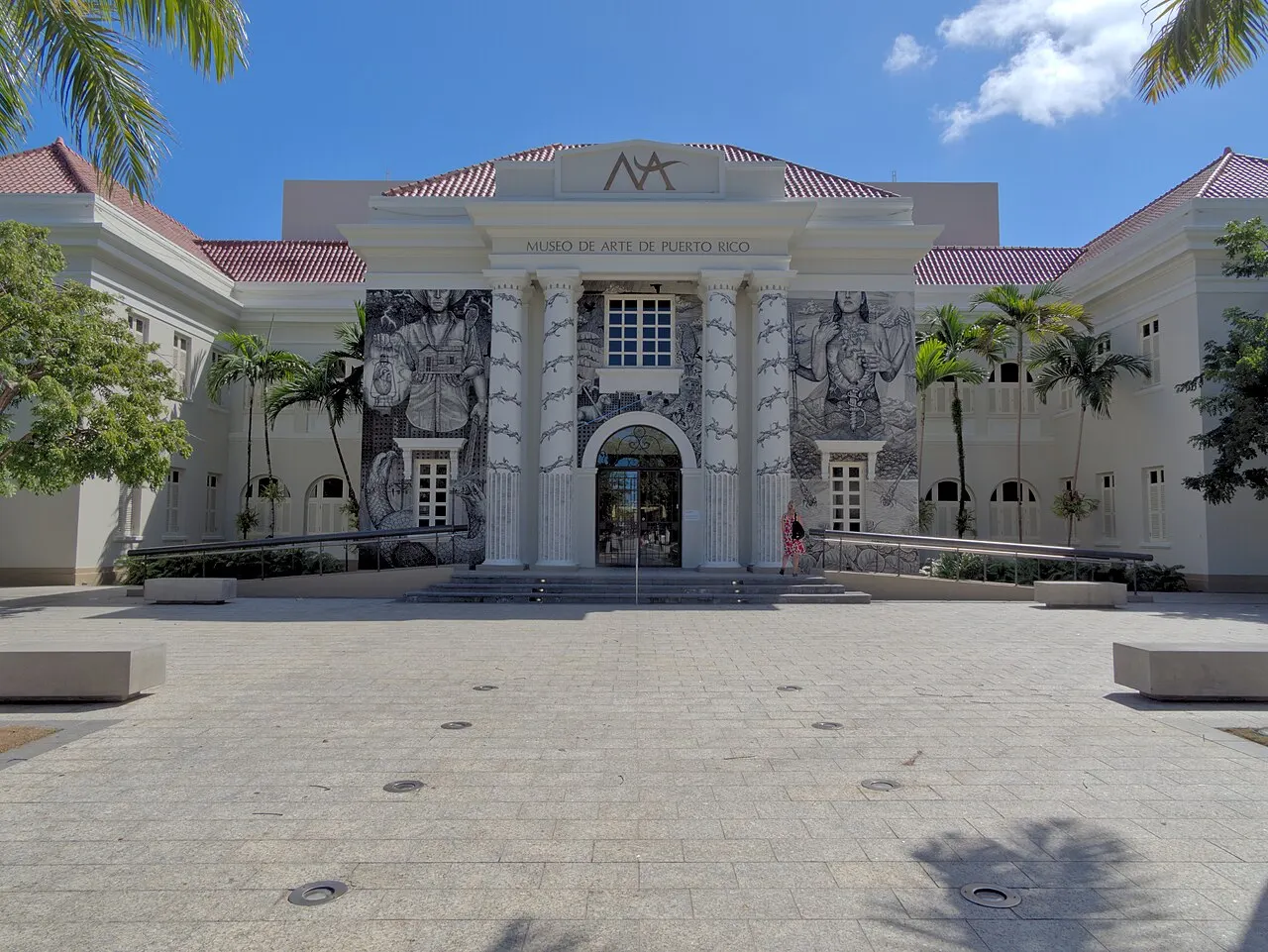 Fachada branca neoclássica do Museo de Arte de Puerto Rico, com colunas, painéis em preto e branco e telhado vermelho, diante de uma praça pavimentada