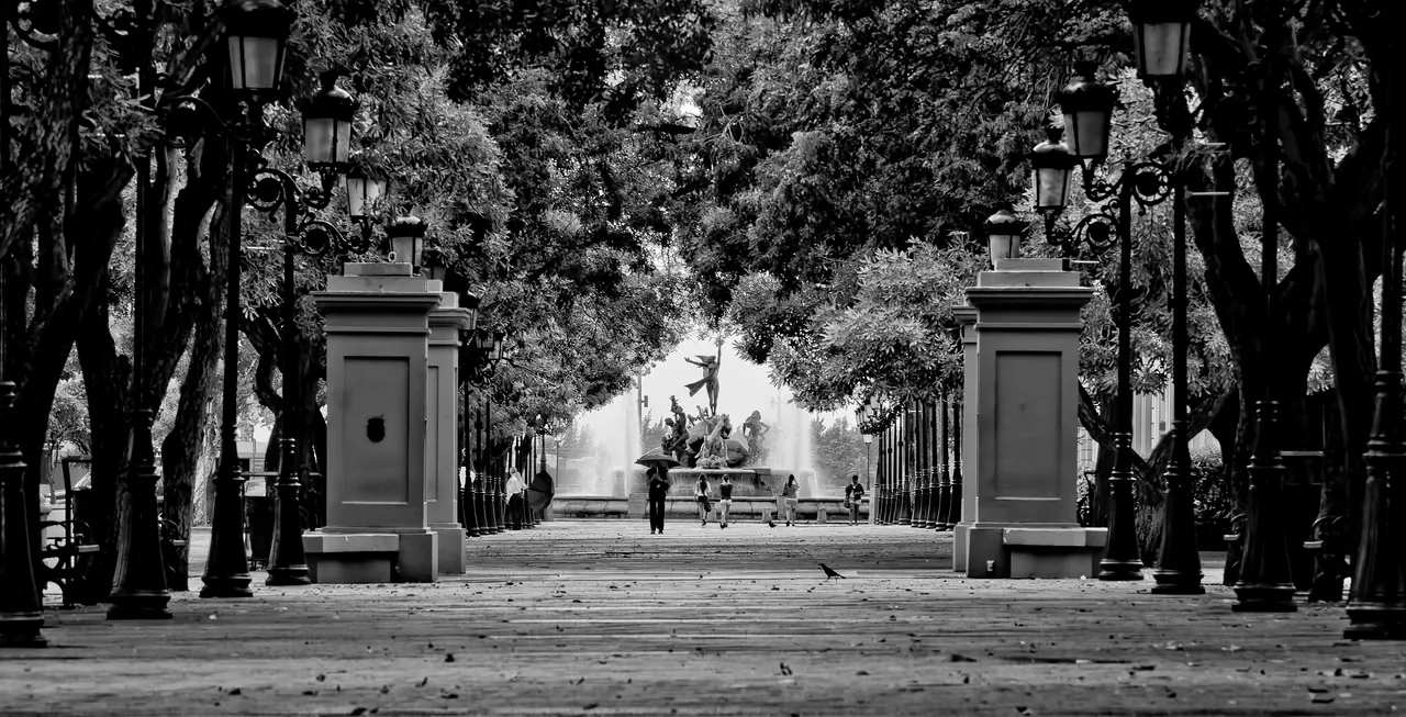 Foto em preto e branco da alameda arborizada do Paseo de la Princesa, com postes de ferro e dois pilares de pedra, e uma fonte com esculturas ao fundo