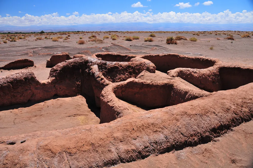 Os muros baixos e curvos de barro da Aldeia de Tulor, formando recintos circulares parcialmente cobertos de areia no deserto, sob céu com nuvens.