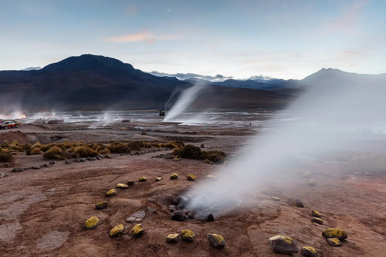 Colunas de vapor saindo do solo nos Géiseres del Tatio, com pedras marcando a área de visita e montanhas escuras ao fundo, na luz do amanhecer.