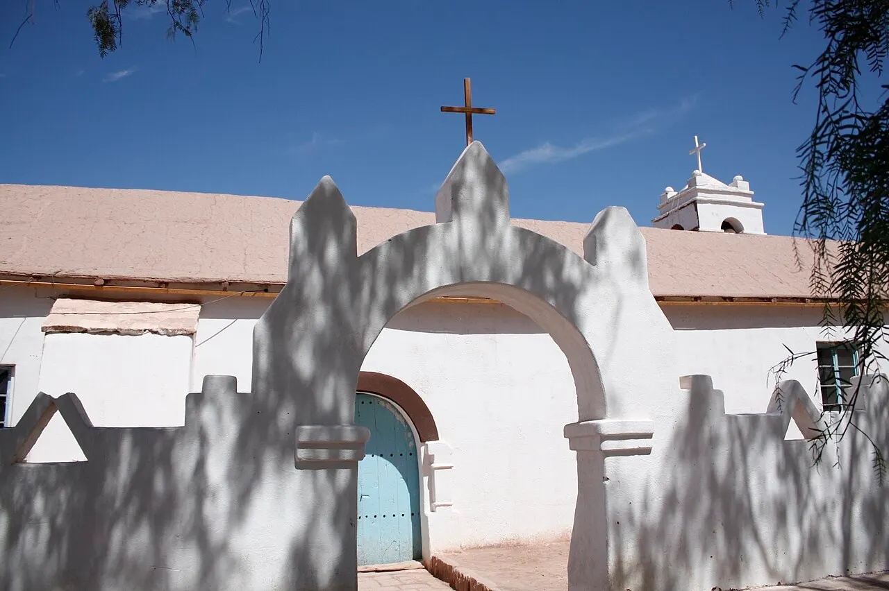 O muro caiado e o portão em arco da igreja de San Pedro de Atacama, com cruzes no topo e o campanário branco ao fundo, sob céu azul.