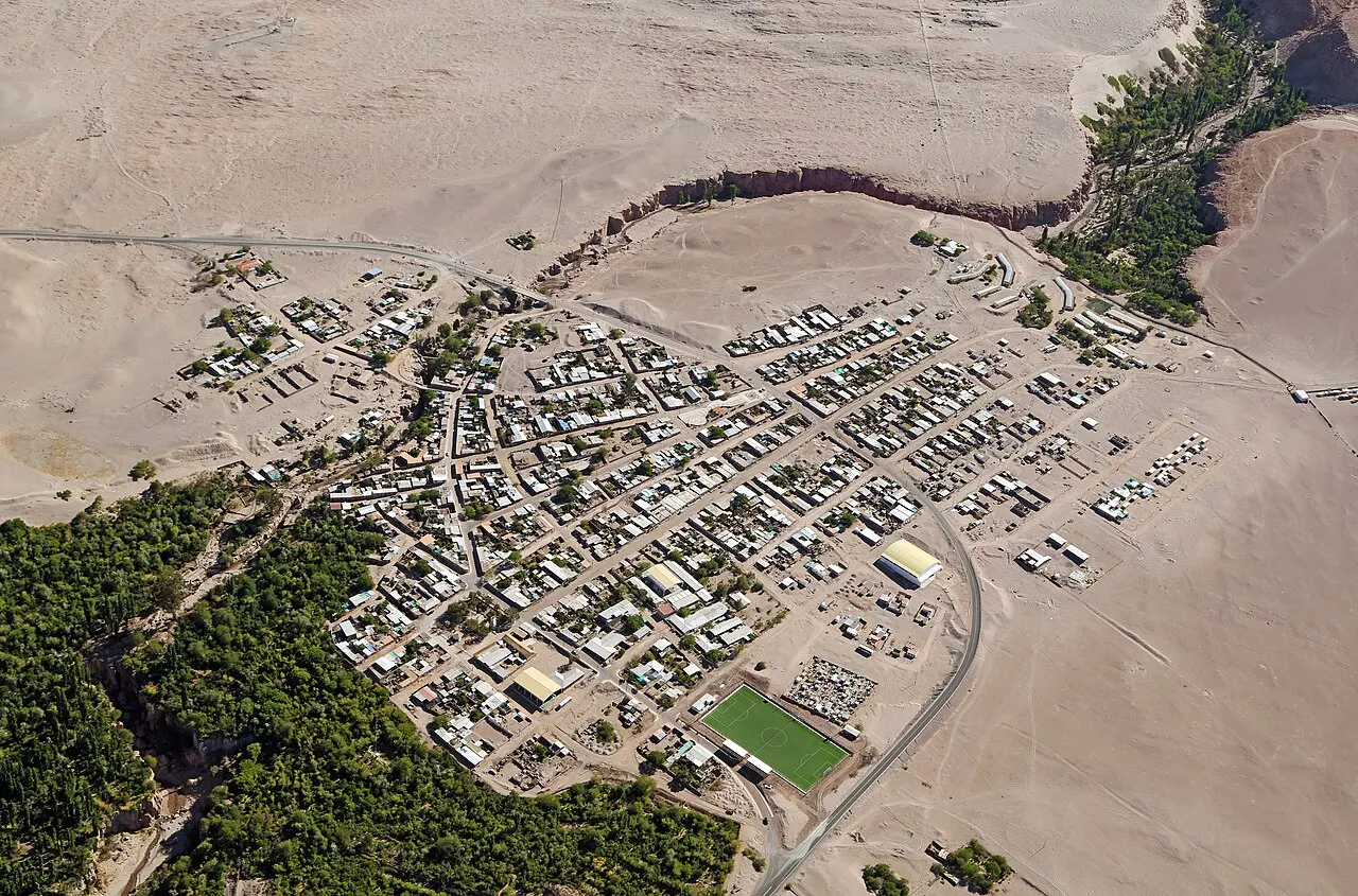 Vista aérea de Toconao, um povoado de casas baixas no deserto, com um campo de futebol verde e a quebrada arborizada ao lado.