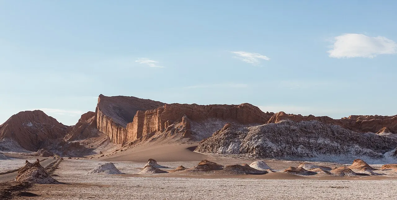 O Anfiteatro do Valle de la Luna, um paredão de rocha avermelhada erguido sobre uma planície clara de sal e argila, sob céu azul.