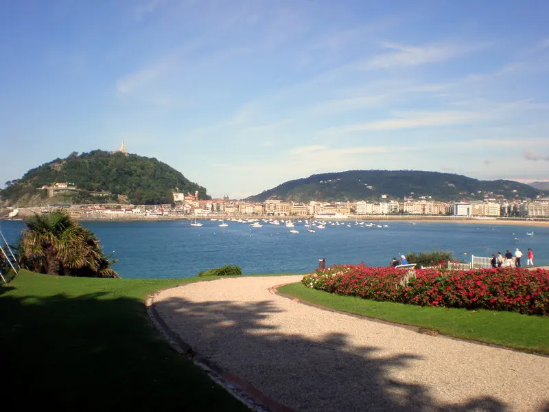 Os jardins do Palácio Miramar com canteiros de flores vermelhas e um caminho, e a baía da Concha com o Monte Urgull ao fundo.
