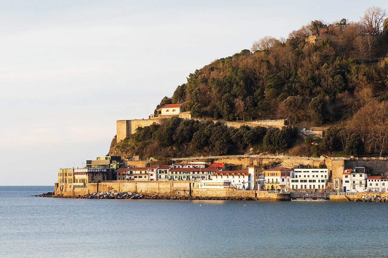 O Monte Urgull coberto de mata com as fortificações no alto e as casas do porto embaixo, na luz do fim da tarde.