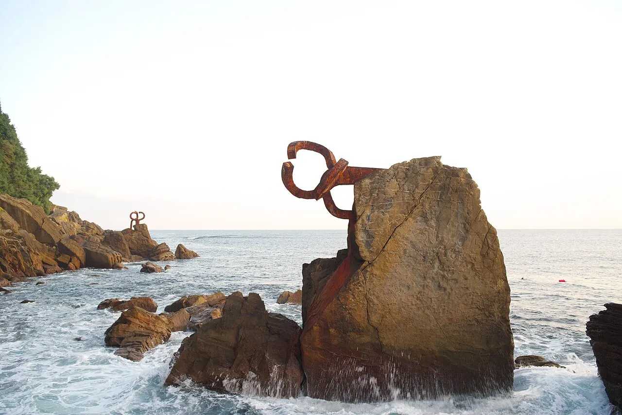 Uma das garras de ferro enferrujado do Peine del Viento presa a uma rocha no mar, com ondas e outra peça ao fundo.