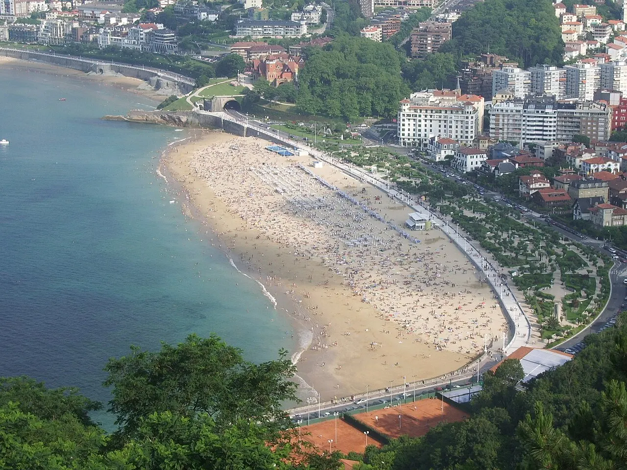 A praia de Ondarreta vista do alto do Monte Igueldo, cheia de banhistas, com o passeio arborizado e os prédios atrás.