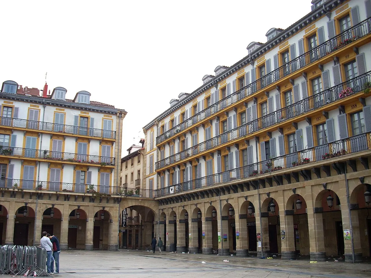 A Plaza de la Constitución cercada de prédios com arcadas no térreo e fileiras de sacadas de ferro, num dia nublado.