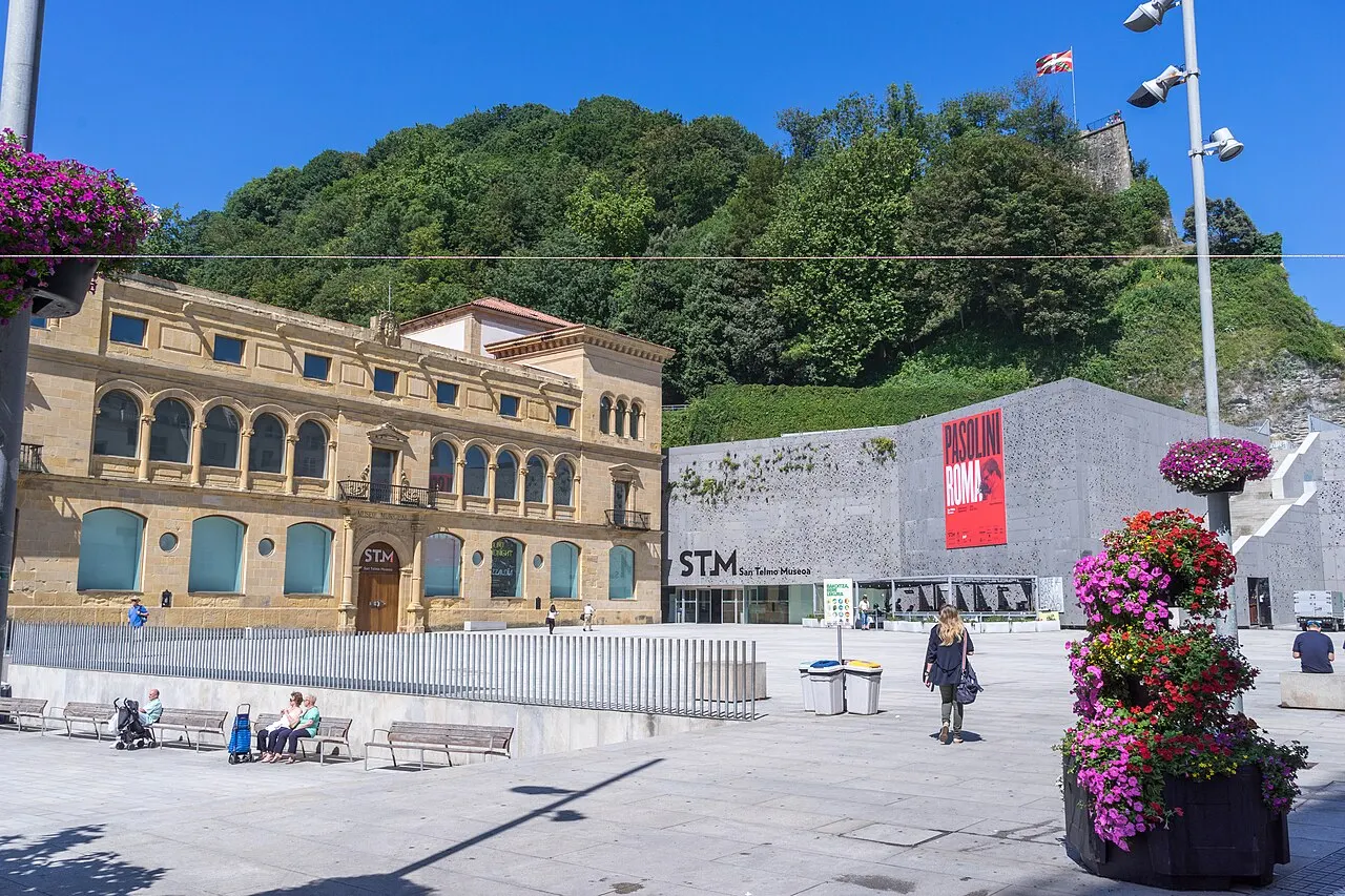 O antigo convento de pedra do San Telmo Museoa e o anexo moderno cinza ao lado, ao pé do Monte Urgull, com uma praça e flores.