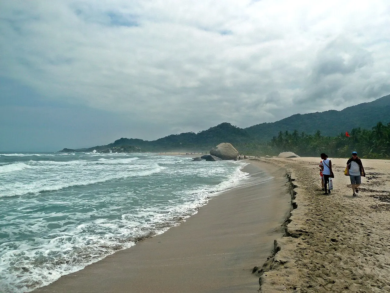 Praia longa de areia escura com ondas fortes quebrando, duas pessoas caminhando na areia e morros com mata e coqueiros sob céu nublado.