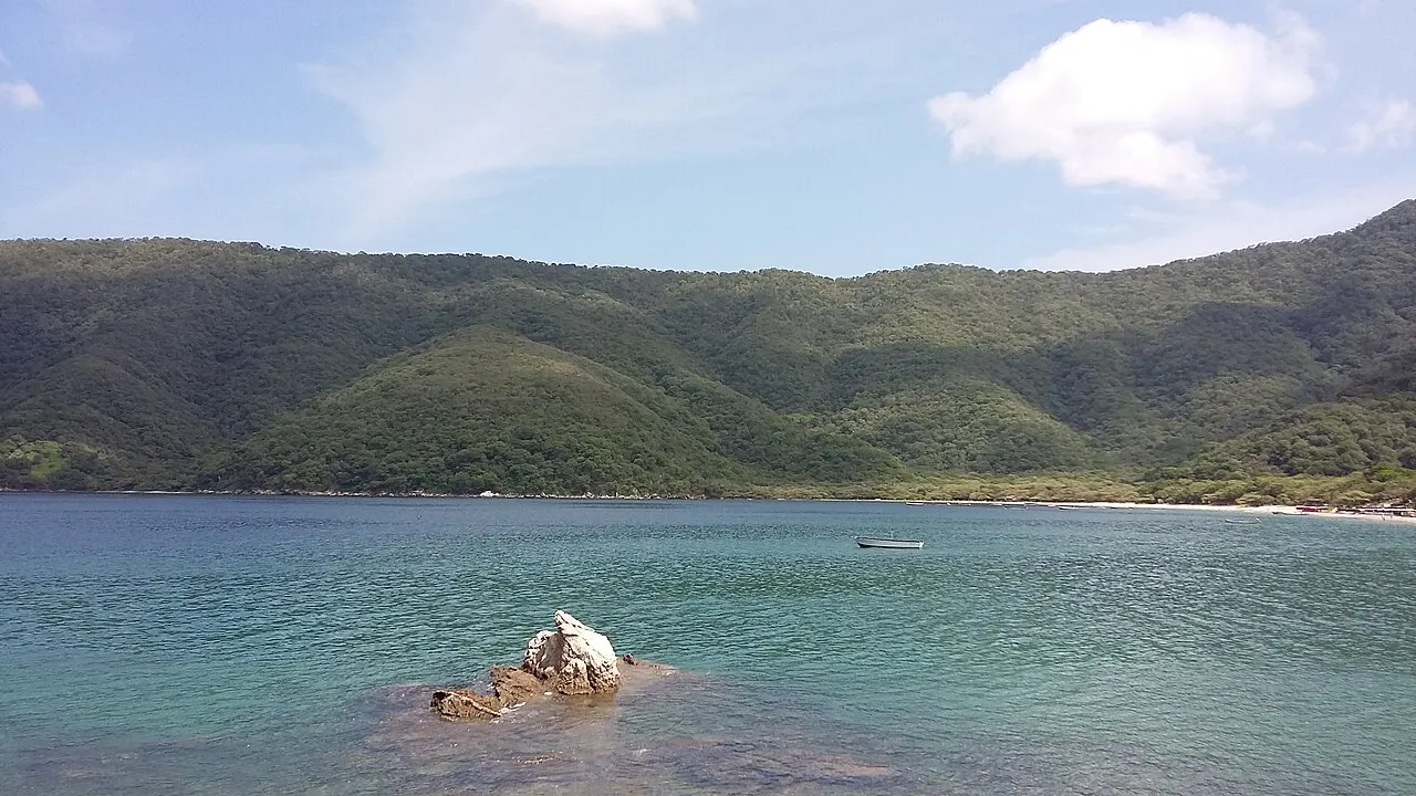 Baía de água calma e esverdeada com uma pedra clara à flor da água em primeiro plano, um barco pequeno ao longe e morros cobertos de mata em volta.