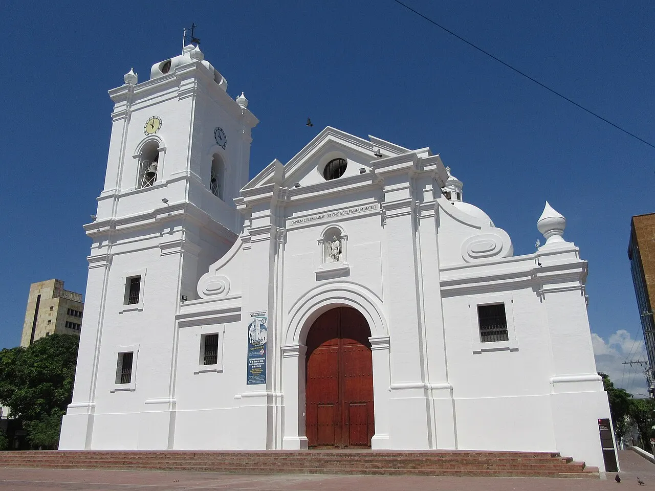 Igreja toda branca com torre de sino e relógio à esquerda, frontão curvo e porta de madeira vermelha em arco, sob céu azul.