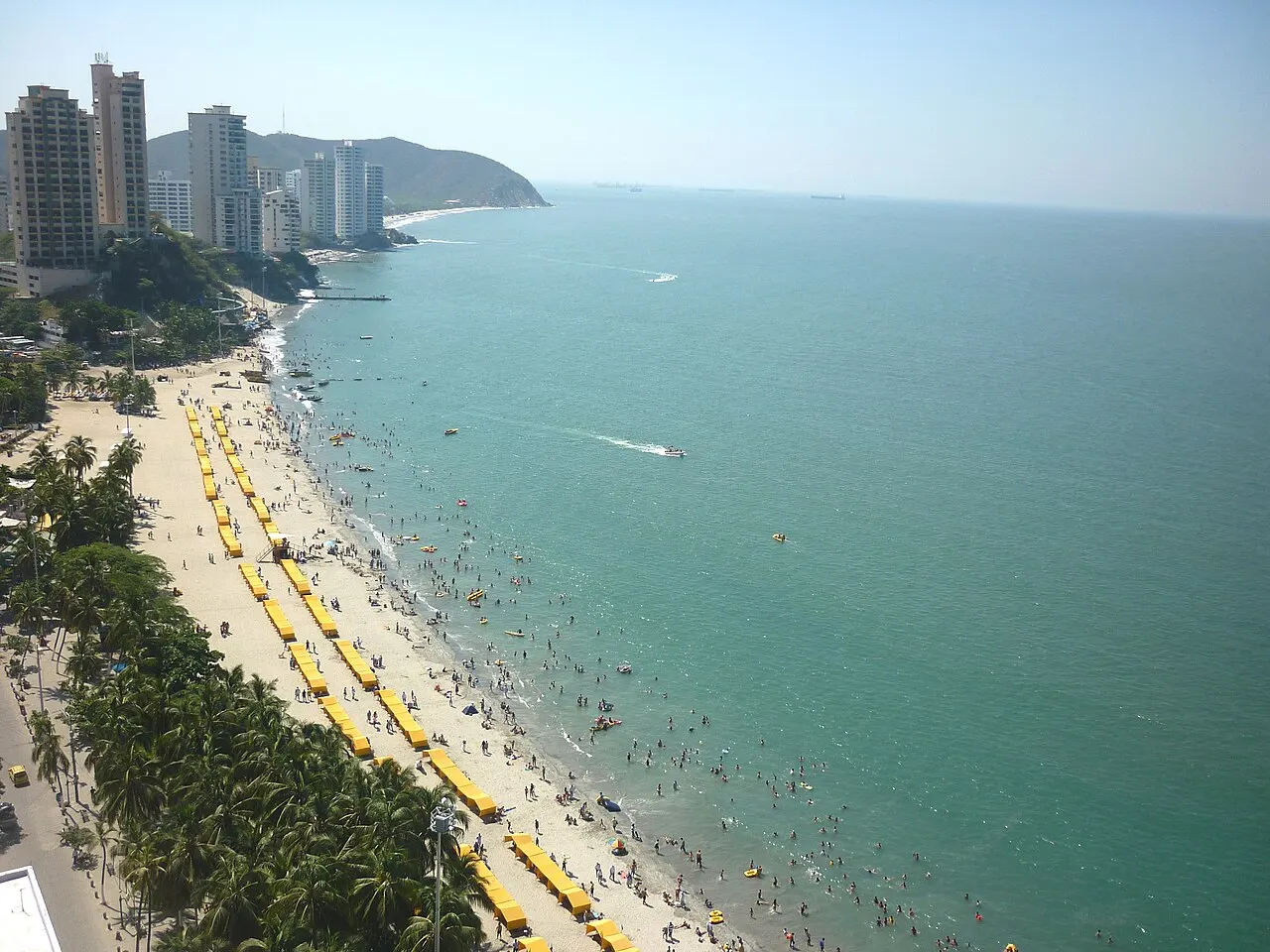 Praia longa e cheia de banhistas, com fileiras de guarda-sóis amarelos, coqueiros na orla, prédios altos à esquerda e mar verde-azulado, vista do alto.