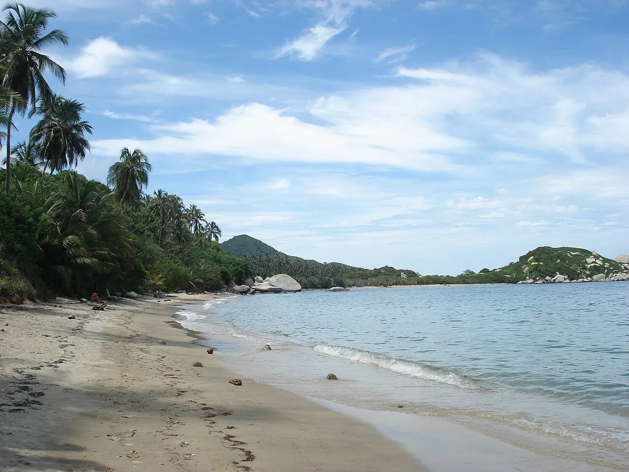Praia de areia clara com água calma, coqueiros e mata à esquerda e grandes pedras arredondadas na ponta da enseada.