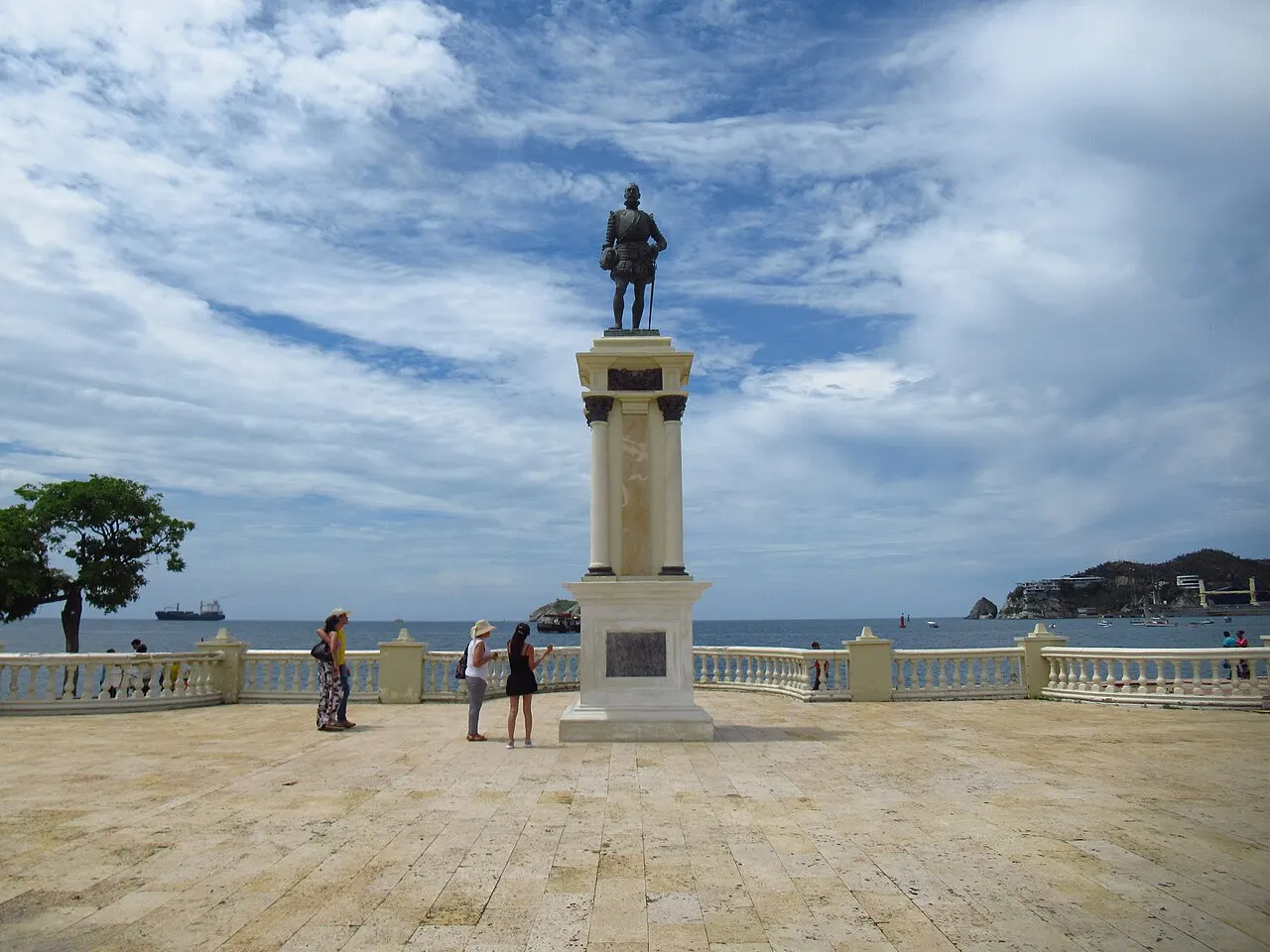 Estátua de bronze de um homem de armadura sobre um pedestal claro, num terraço com balaustrada branca à beira da baía, com navios e um morro ao fundo.