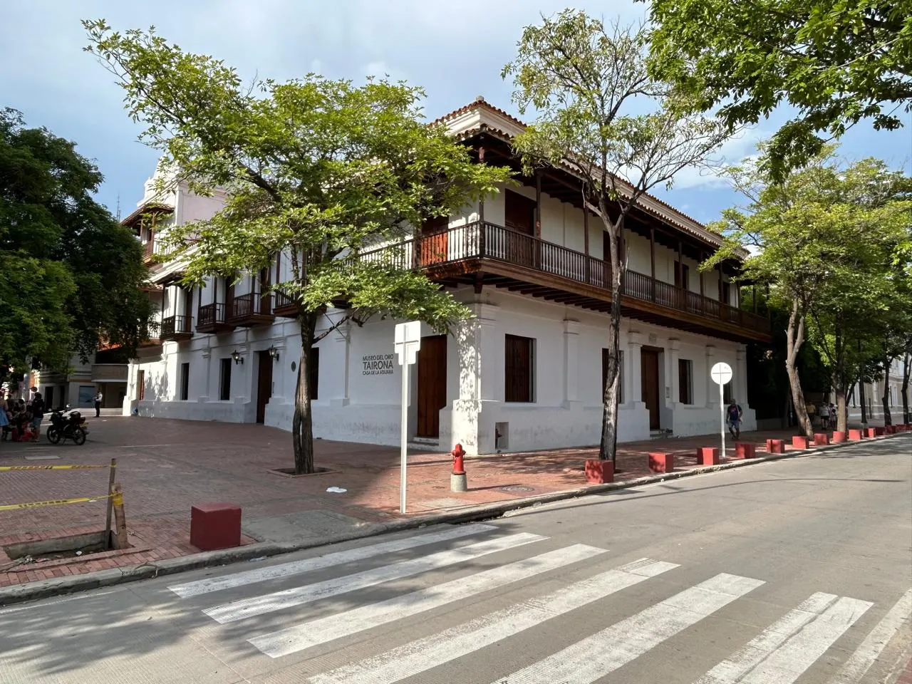 Casarão colonial branco de dois andares com varanda de madeira escura no andar de cima, numa esquina com árvores e faixa de pedestres.