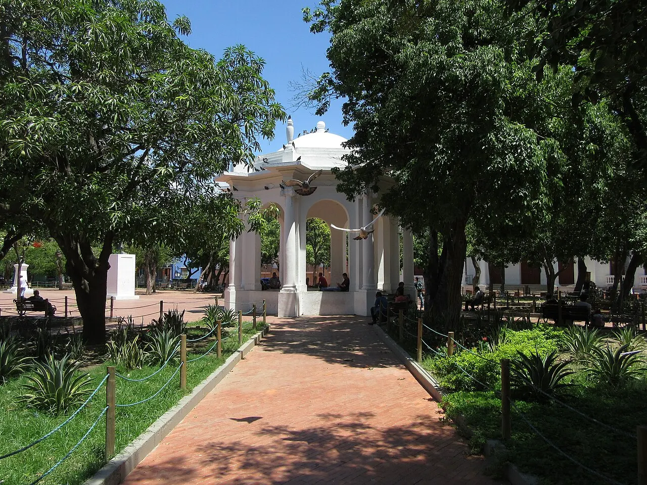 Coreto branco de arcos e cúpula no meio de uma praça arborizada, com um caminho de tijolo cercado de canteiros e pombos voando.