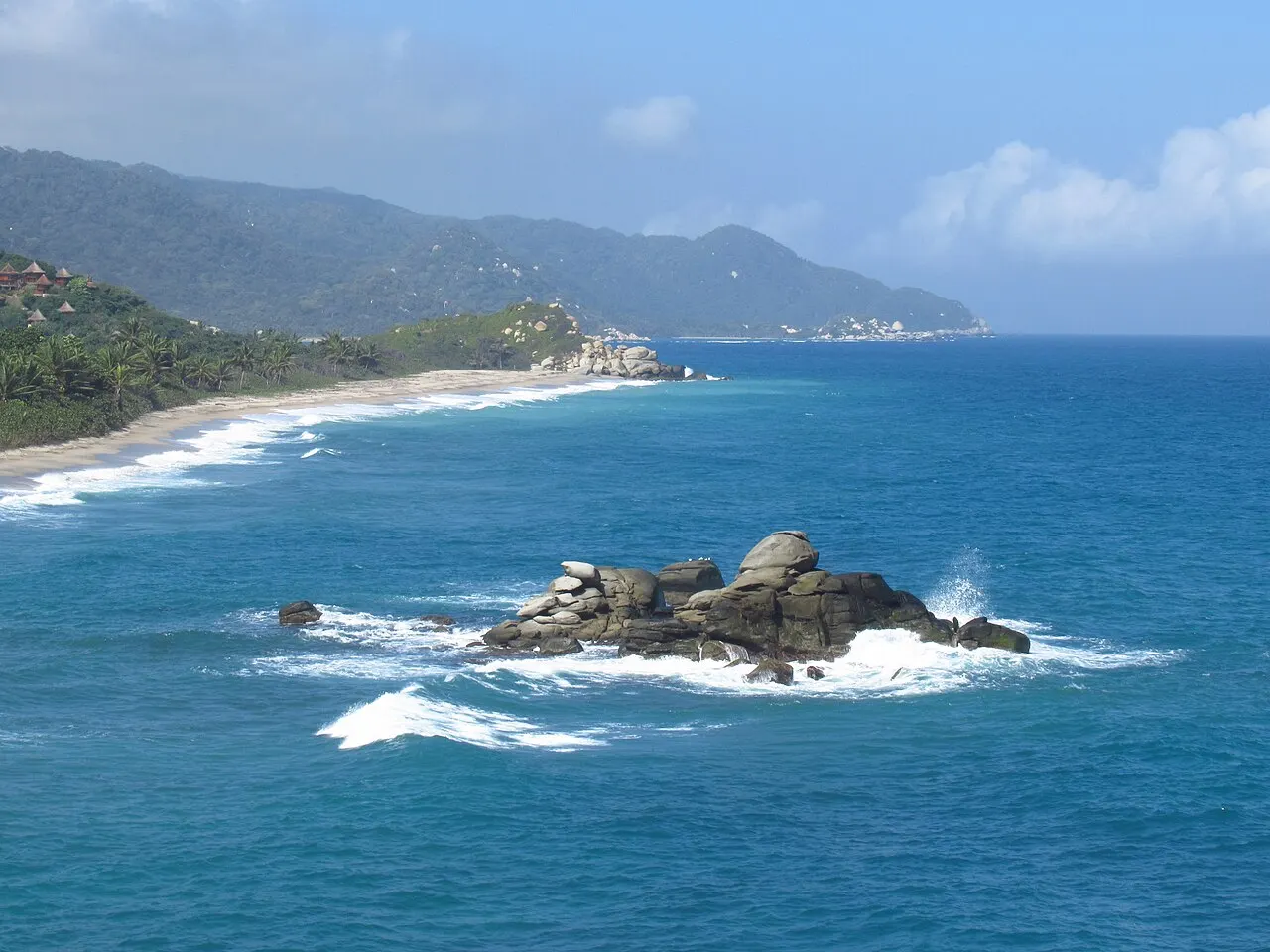 Mar azul com um rochedo batido pelas ondas em primeiro plano, uma praia longa de areia clara e morros cobertos de mata ao fundo.
