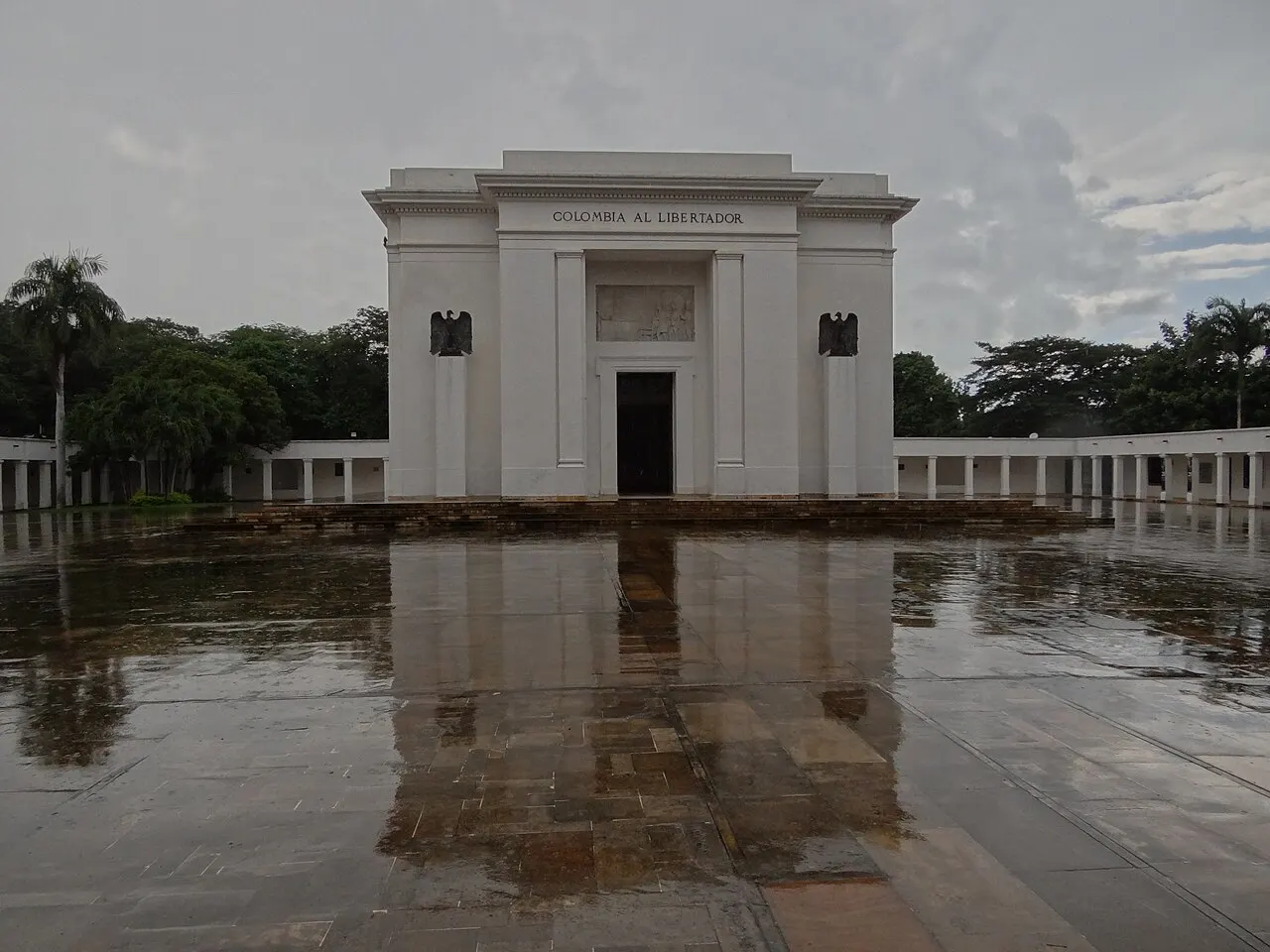Monumento branco de linhas retas com a inscrição «Colombia al Libertador», duas águias na fachada e colunatas dos lados, diante de uma praça molhada de chuva.