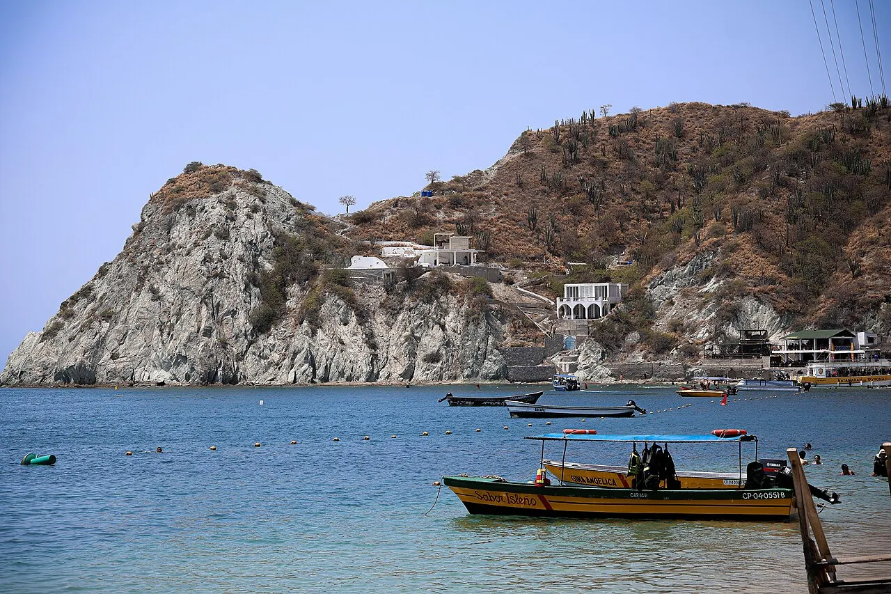 Lanchas de pesca na água azul da baía, diante de um morro de pedra clara e vegetação seca, com casas brancas na encosta.