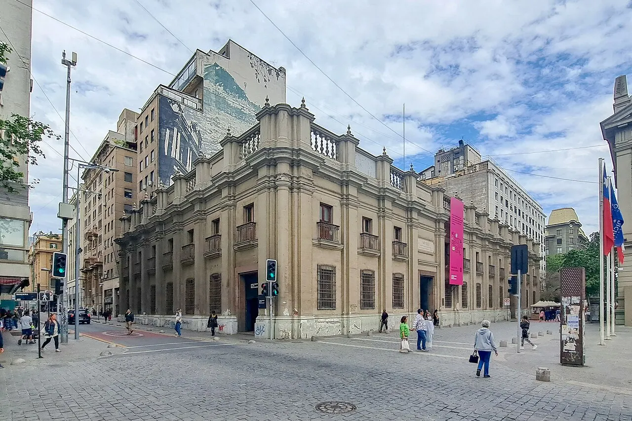 Fachada neoclássica de esquina do Museo Chileno de Arte Precolombino, com balaustrada no telhado, uma bandeira do Chile e pessoas atravessando a rua.