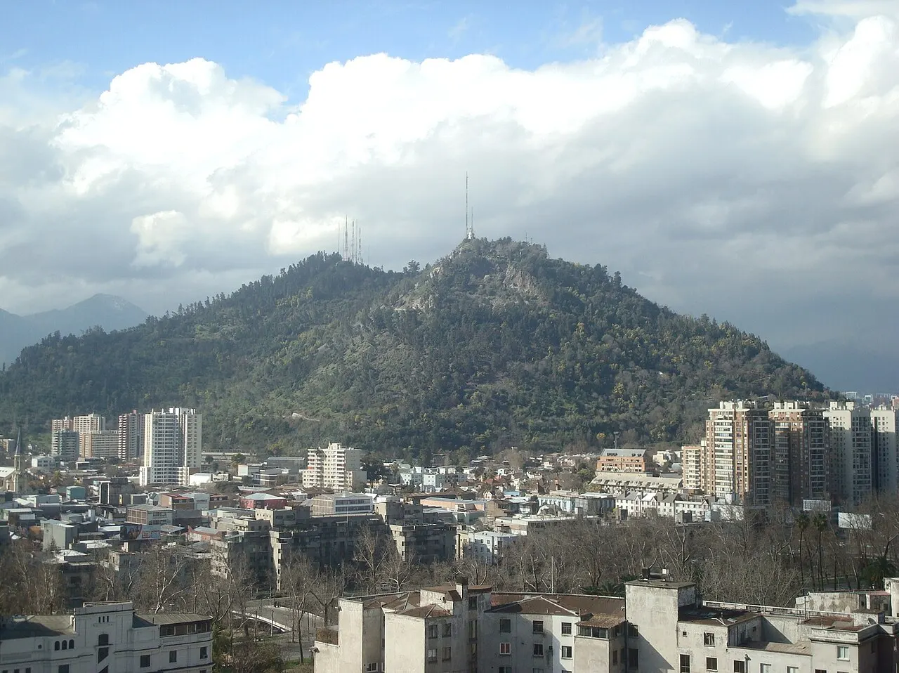 O Cerro San Cristóbal, coberto de vegetação, com prédios da cidade na base e nuvens no céu.