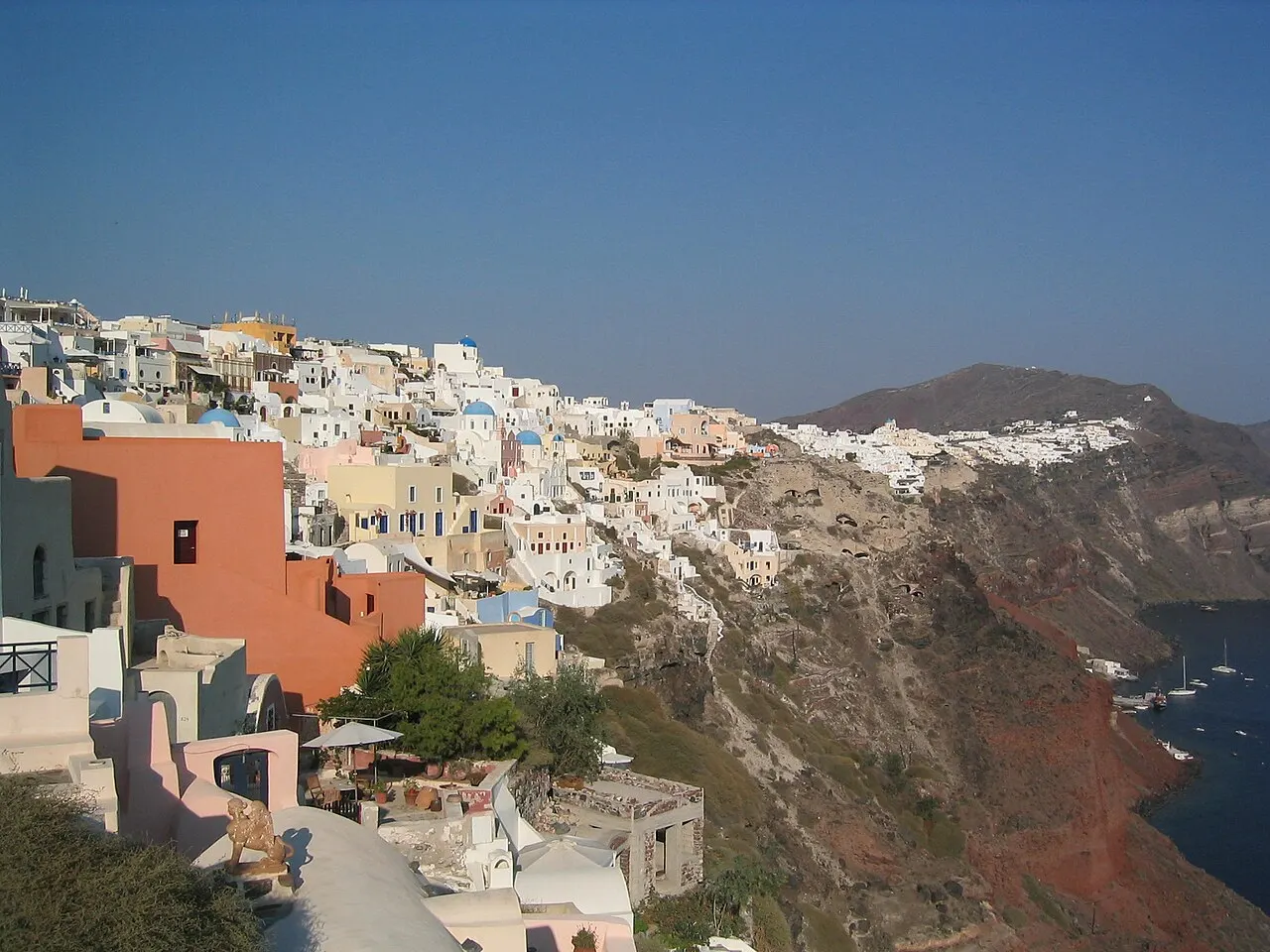 Casas brancas e cúpulas azuis de Oia escalonadas na borda da caldera de Santorini, com a rocha vermelha e escura caindo até o mar azul à direita.