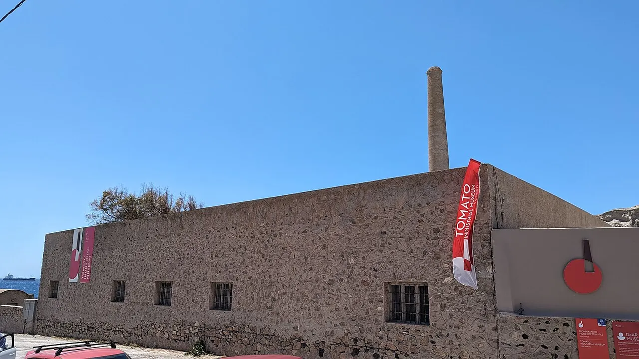 Prédio de pedra da antiga fábrica do Museu Industrial do Tomate, com chaminé alta e faixa vermelha, sob céu azul.