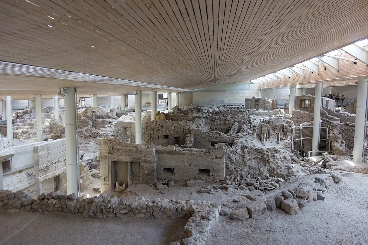 Ruínas de pedra do sítio pré-histórico de Akrotiri, com paredes e portas antigas preservadas sob uma grande cobertura moderna de madeira e vidro.