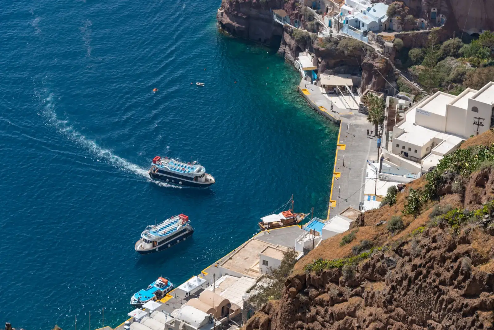 O porto velho de Fira visto do alto da falésia: cais estreito de concreto ao pé da rocha vermelha, dois barcos de excursão manobrando na água azul-escura e as casas brancas da borda lá em cima.