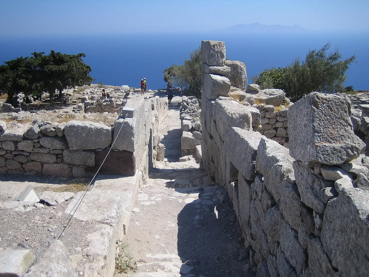 Ruínas de muros de pedra da Thera Antiga ao longo de uma rua estreita no alto do monte Mesa Vouno, com o mar Egeu e uma ilha ao fundo.