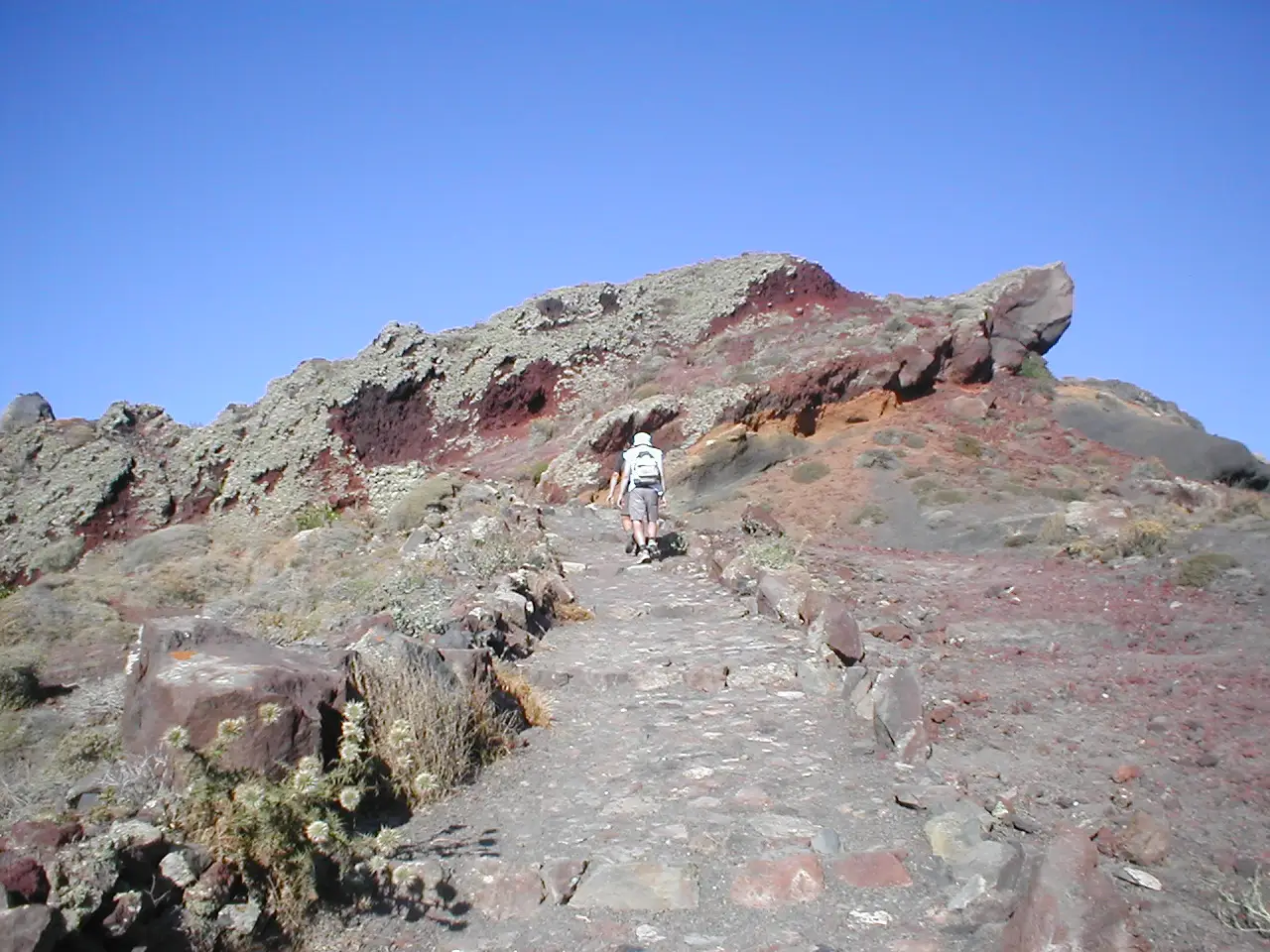 Um caminho estreito de pedra sobre uma crista avermelhada, com o mar à esquerda e uma pessoa caminhando à frente.