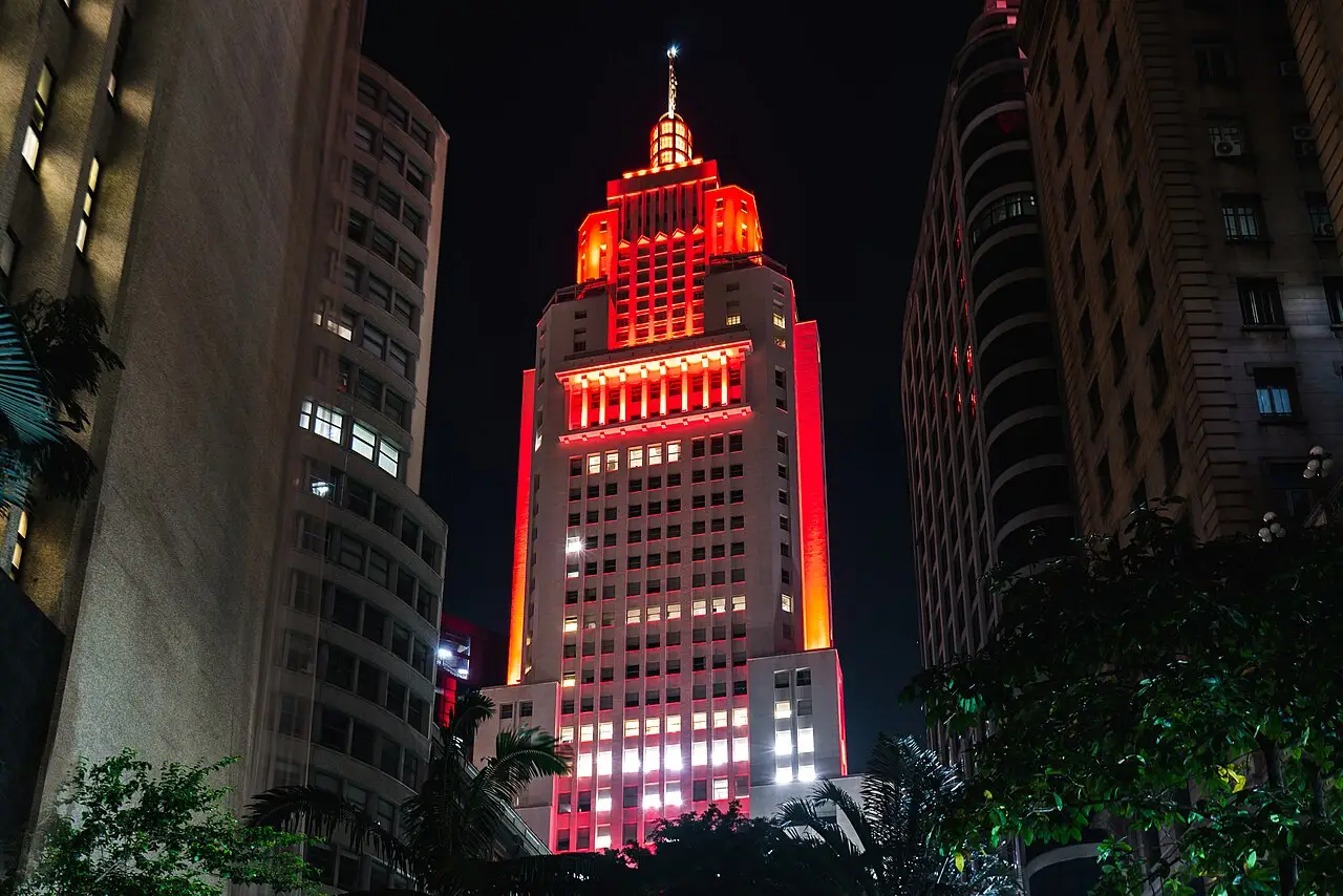 Torre alta iluminada de vermelho e dourado, com o topo em degraus, entre prédios.