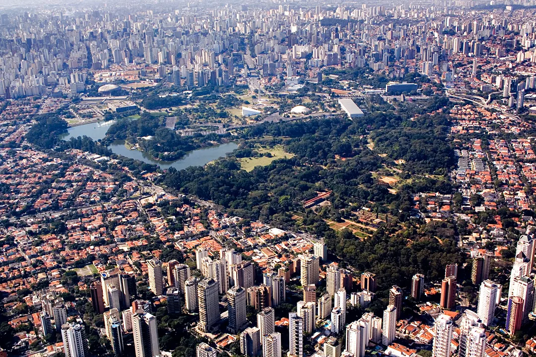 Vista aérea do Parque Ibirapuera, com um lago cercado de árvores e gramados, prédios de museus dentro do parque e a cidade densa ao redor.
