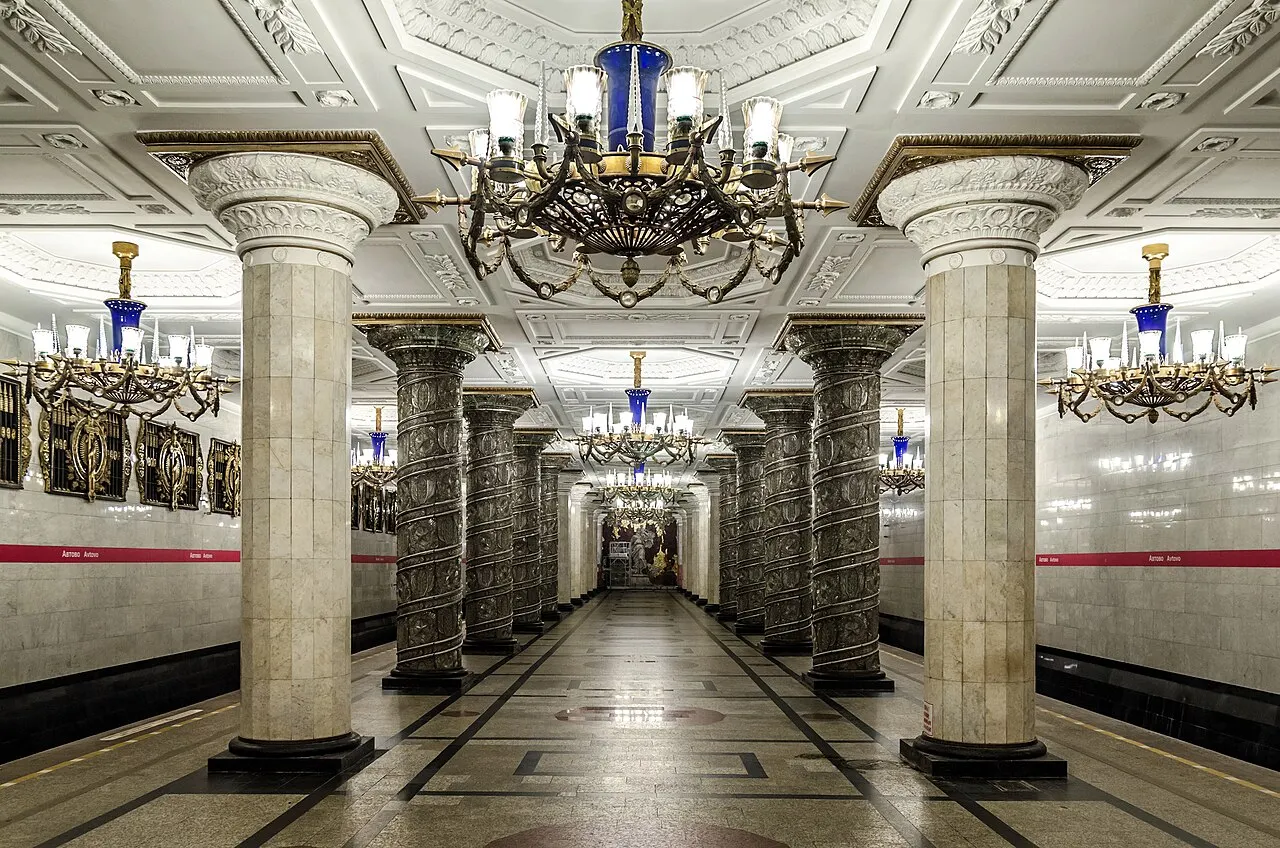 Salão da estação Avtovo do metrô, com colunas de pedra clara, lustres e piso brilhante em perspectiva central.
