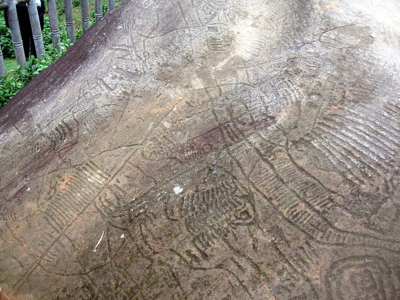 Superfície de uma grande pedra cinza coberta de gravuras: linhas paralelas, espirais e figuras geométricas riscadas na rocha, com uma cerca de madeira no canto.