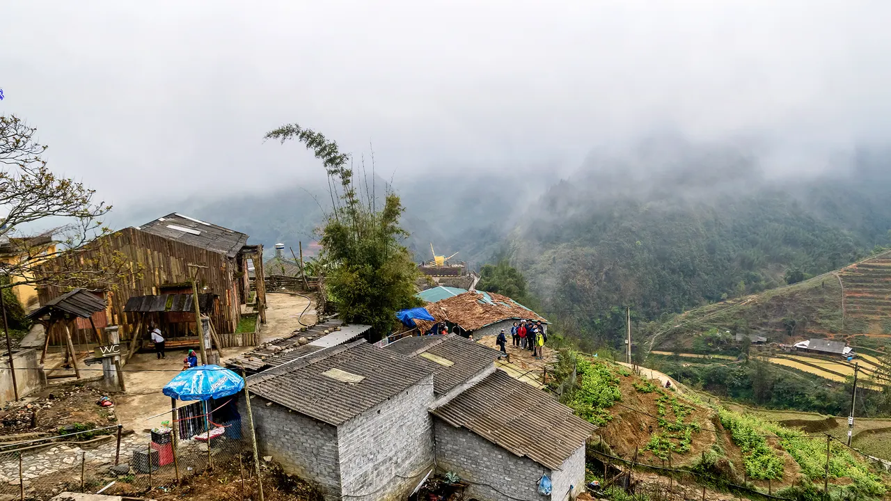 Casas de madeira e de blocos com telhados de chapa na encosta da aldeia de Cát Cát, com terraços de arroz abaixo e montanhas cobertas de névoa ao fundo.