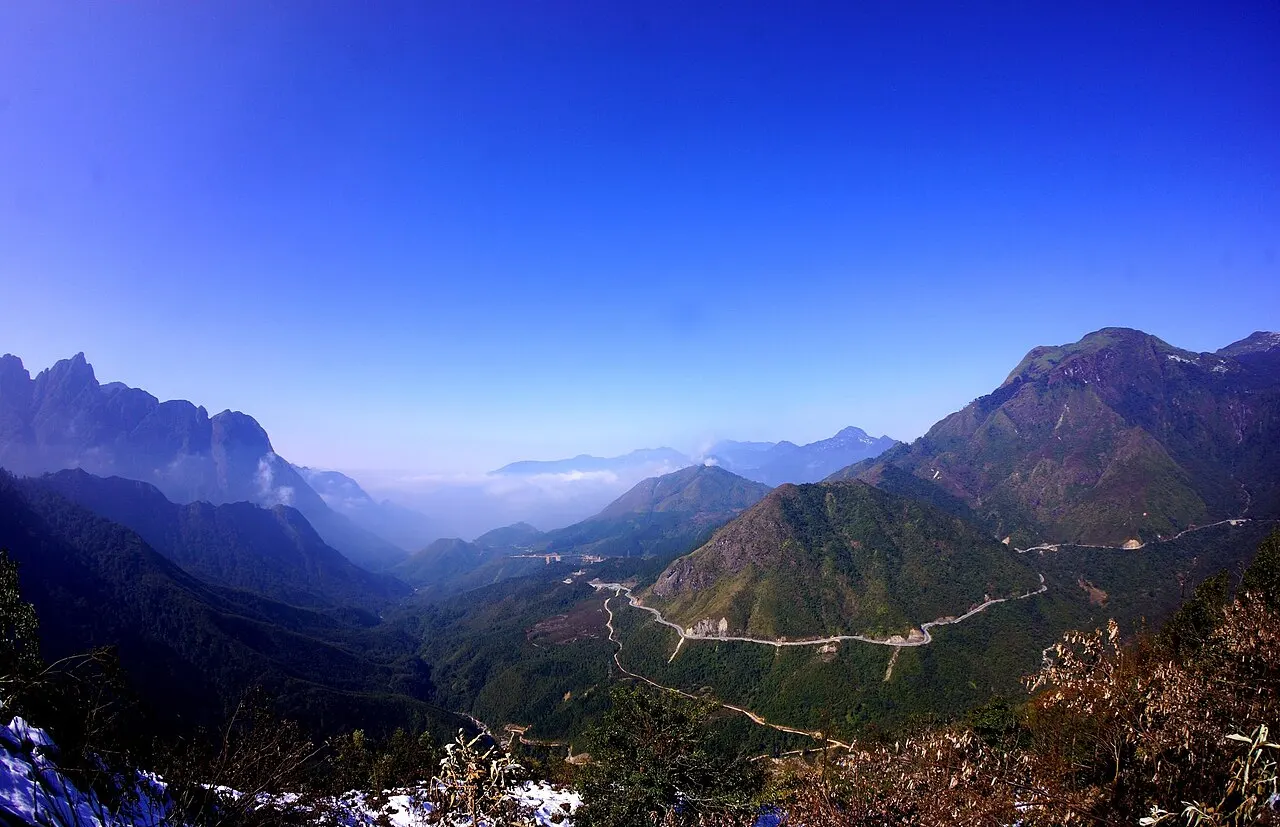 The Ô Quy Hồ pass road winding through a valley between high mountains under a blue sky, with dry vegetation and a little frost in the foreground.