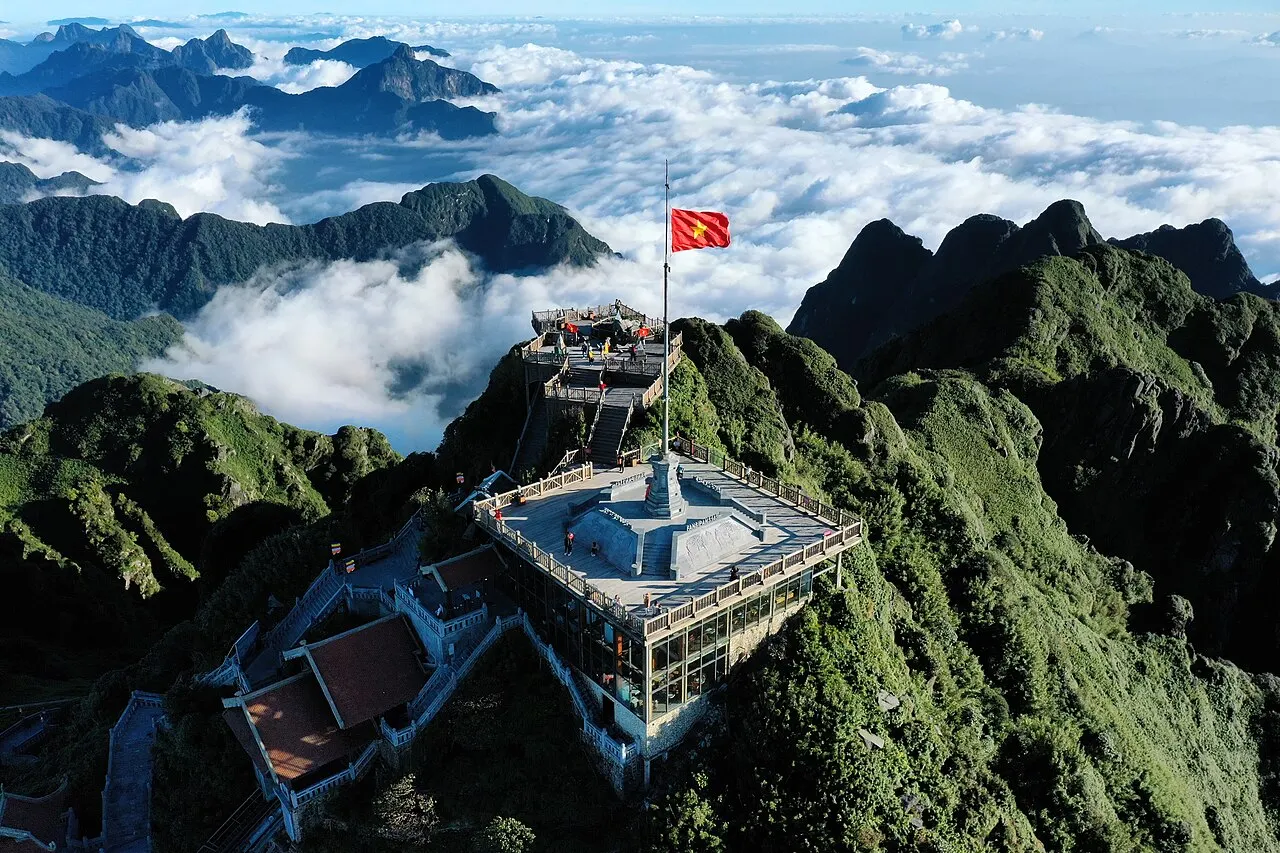 Aerial view of the Fansipan summit: the platform with the summit marker and a pole flying the Vietnamese flag on the green ridge, with peaks and a sea of clouds behind.