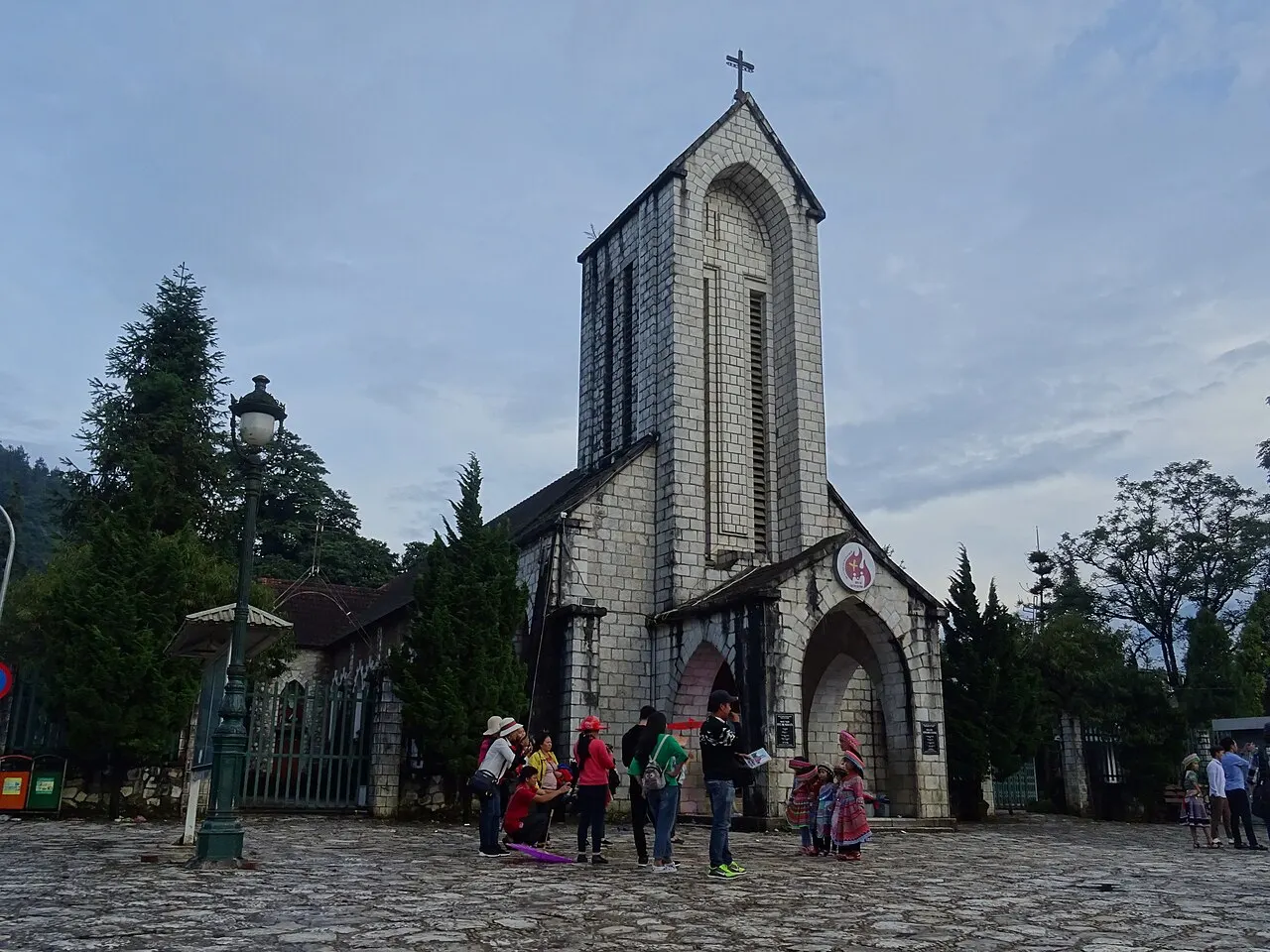 Sa Pa's grey stone church, with a tall tower and a cross on top, seen from the cobbled square, with pine trees beside it and people in front of the entrance.