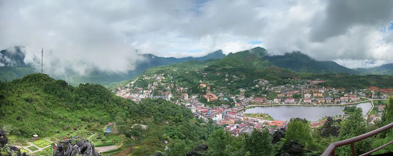 Vista do alto do monte Hàm Rồng: a cidade de Sa Pa espalhada no vale em volta do lago, com encostas verdes, um corrimão no primeiro plano e nuvens sobre as montanhas.