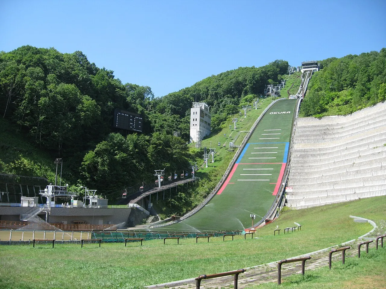 A green-surfaced ski jump runs down a wooded slope to a lawn, with white stands on the right, a chairlift and a white tower beside it, under a blue sky.