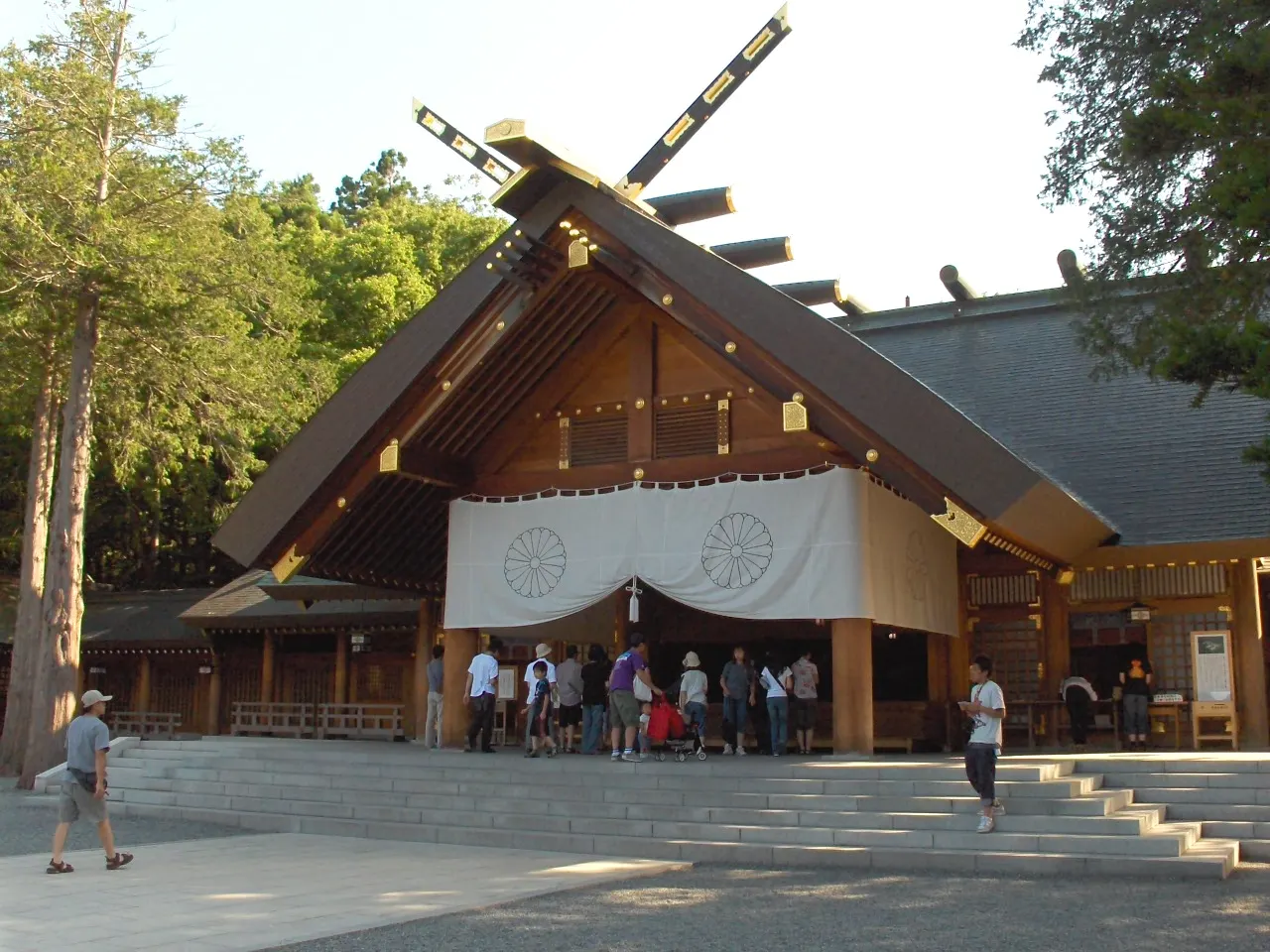 Pabellón de santuario de madera con tejado inclinado y vigas cruzadas en lo alto, una cortina blanca con dos emblemas en la entrada y visitantes en la escalinata de piedra, entre árboles altos.